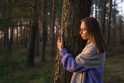 Loving nature concept. relaxed woman hugging tree trunk in autumn forest with closed eyes at sunset