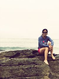 Portrait of smiling young woman sitting on rock at shore against clear sky