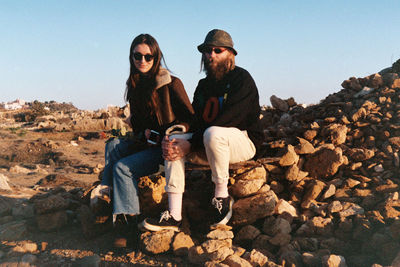 Portrait of young man and woman sitting on rock against sky