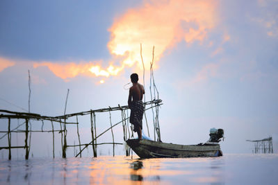 Man standing on fishing boat in sea against sky during sunset