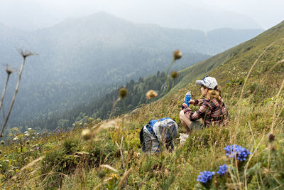 Young woman sitting on meadow in mountain by backpack drinking water from bottle , hiking landscape 