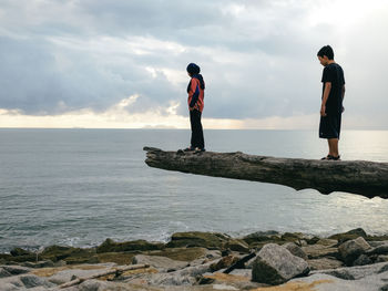 Rear view of a boy and a girl standing on drift  wood by sea against cloudy sky