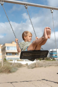 A happy boy on a swing on a sandy beach against the backdrop of apartment buildings and a stormy sky