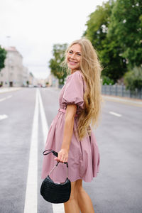 Young woman in a pink dress holding a black handbag, smiling over her shoulder while standing