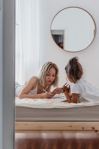 Caucasian mom and hispanic daughter painting while they are lying in the bed.