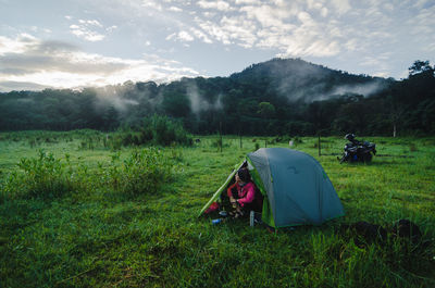 Woman sitting by tent on grass against mountains at sunset