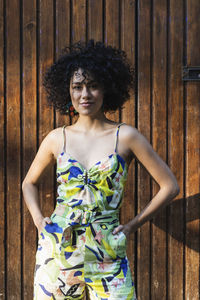 Young woman with hands in pockets standing against wooden wall