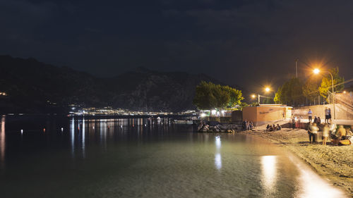 Illuminated street by river against sky at night