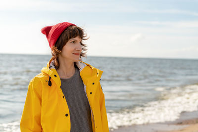 Waist portrait of mature woman in bright yellow cloak and red hat on shore of north sea on cold