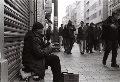 Man playing musical instrument by people walking on footpath in city