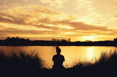 Rear view of silhouette man sitting by lake against sky during sunset