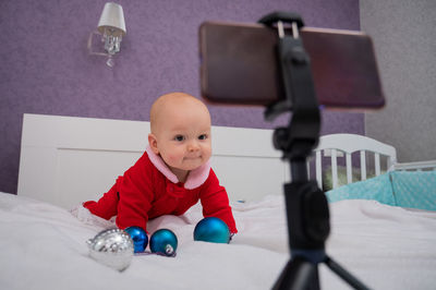 A baby boy dressed in a santa suit sits on the bed and looks at the sick phone. christmas greetings
