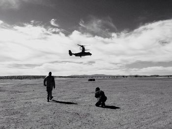 Rear view of army soldiers on landscape with helicopter in mid-air against sky