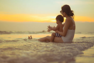 Woman sitting on shore at beach against sky during sunset