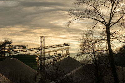 Bare trees by factory against sky during sunset