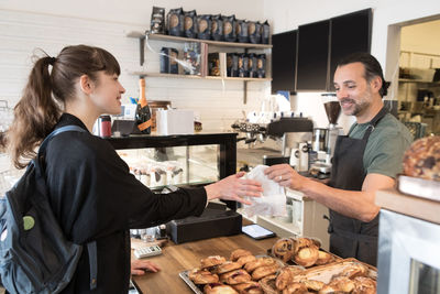 Smiling mature male owner serving food package to female customer at checkout in bakery