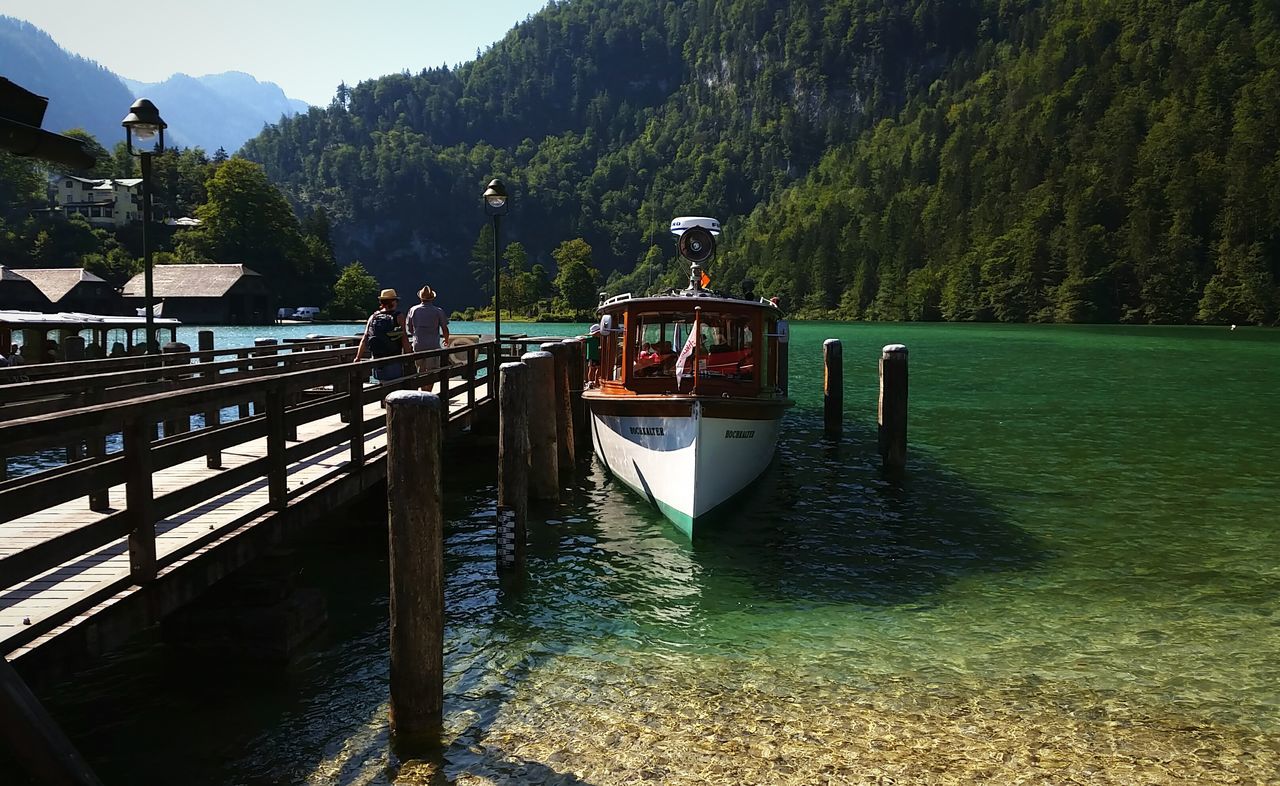 tree, water, built structure, mountain, river, architecture, transportation, tranquility, nature, railing, incidental people, tranquil scene, canal, bridge - man made structure, day, nautical vessel, building exterior, outdoors, connection, scenics