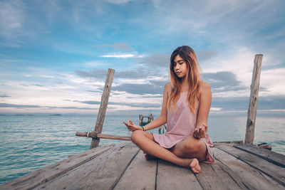 Full length of woman meditating while sitting on pier against sea and sky