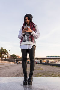 Low angle of young african american female in trendy outfit standing on paved promenade and surfing internet on smartphone in evening in seville