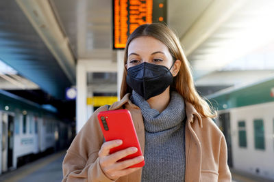 Young woman with black medical mask kn95 ffp2 using telephone in the train station with timetables 