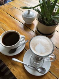 A latte and a cappuccino with a small bowl of sugar and a plant on a wooden table.