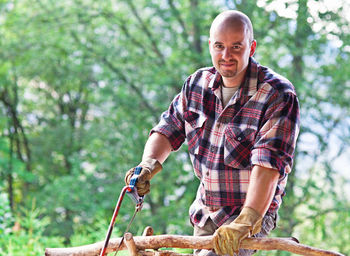 Portrait of man cutting branch against tree