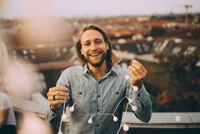 Portrait of cheerful man holding string light while standing on terrace in city