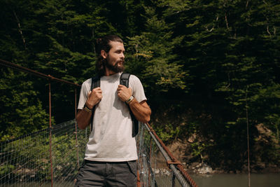 Young man looking away while standing on rope bridge against trees