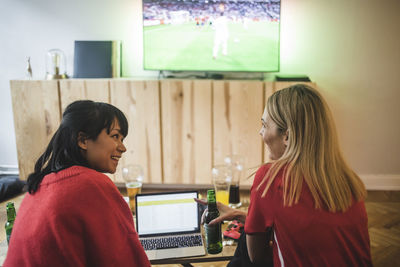 Smiling female friends talking while watching soccer match at home