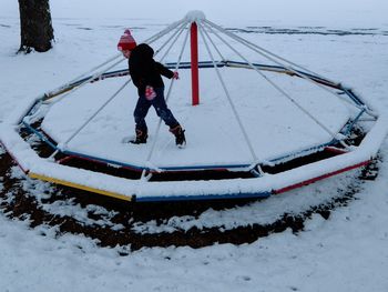 Boy playing on snow covered merry-go-round at park during winter