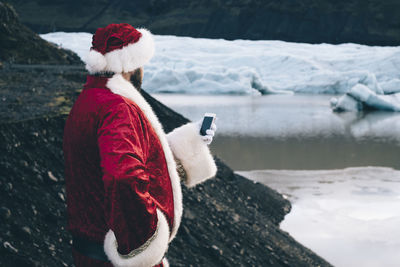 Side view of man wearing santa claus costume using phone by lake during winter