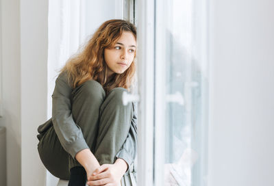 Beautiful sad unhappy teenager girl with curly hair sitting on window sill