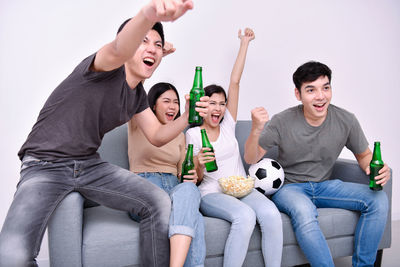 Happy friends watching sports while sitting on sofa against wall