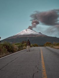Rear view of man on road against smoke emitting from volcano and sky