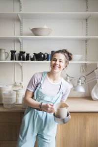 Portrait of smiling young female potter holding pitcher against shelves at workshop