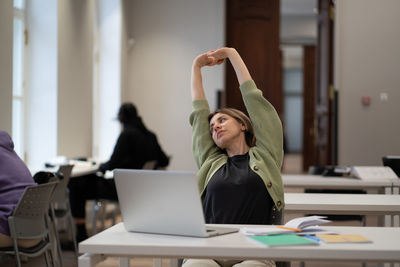 Relaxed middle-aged female stretching hands taking break from work on laptop in library or classroom