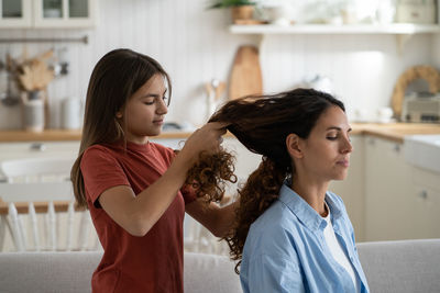Daughter braiding mothers long wavy hair, mom and child enjoying time with each other at home