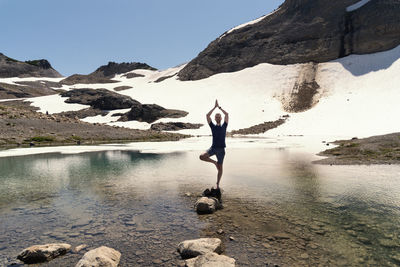 Full length of man doing yoga while standing on rock in lake against mountain
