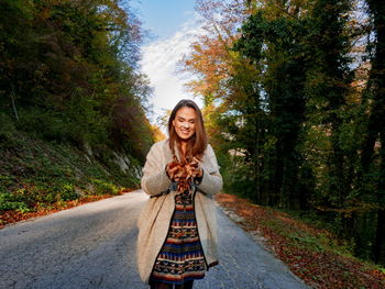 Portrait of smiling young woman standing on road amidst trees