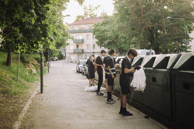 Full length of friends standing with garbage bags by recycling bins