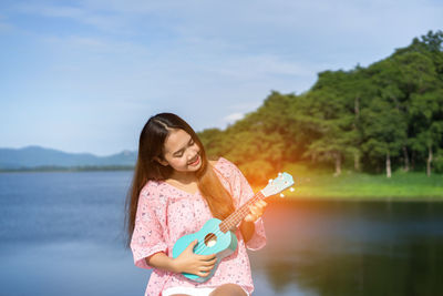 Smiling young woman playing ukulele against lake and sky