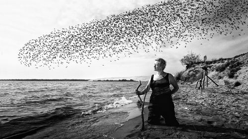 Woman kneeling at beach against birds flying in sky