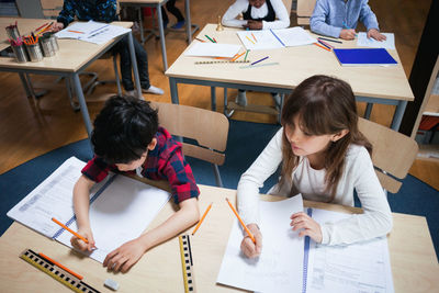 High angle view of students writing on paper while sitting at desk in classroom