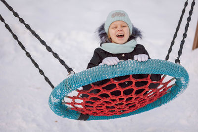 Cheerful girl swings on wicker swing for walk outdoors on frosty winter day in park