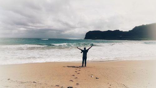 Rear view of man with arms outstretched standing on beach against sky