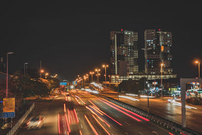 Light trails on road amidst buildings against sky at night