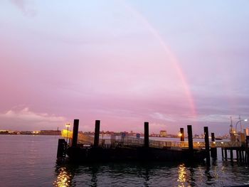 Pier over river against rainbow in sky at sunset