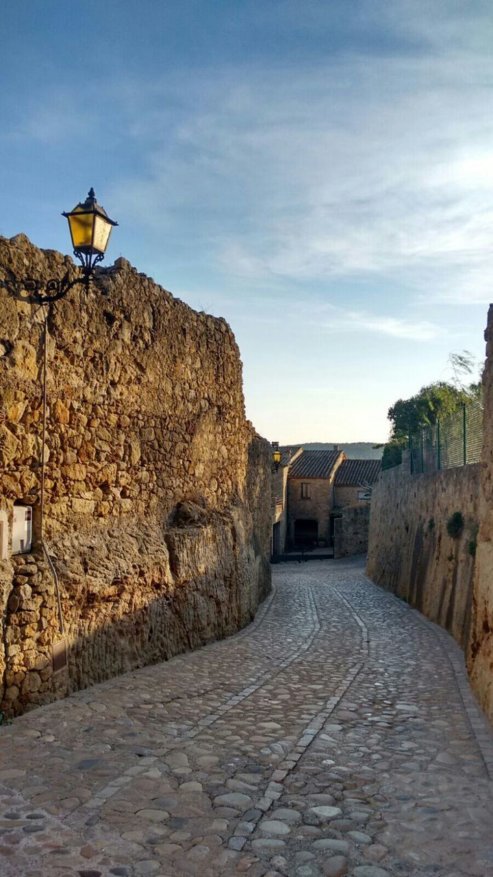 architecture, built structure, building exterior, the way forward, sky, street light, cobblestone, street, cloud - sky, road, footpath, stone wall, house, walkway, lighting equipment, outdoors, day, empty, wall - building feature, cloud