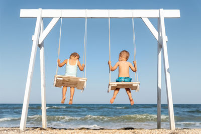 Rear view of siblings sitting on swing st beach