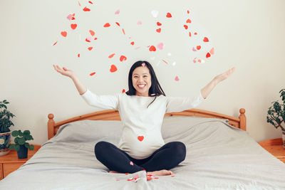 Smiling asian chinese pregnant woman sitting on bed throwing small red pink paper hearts. 